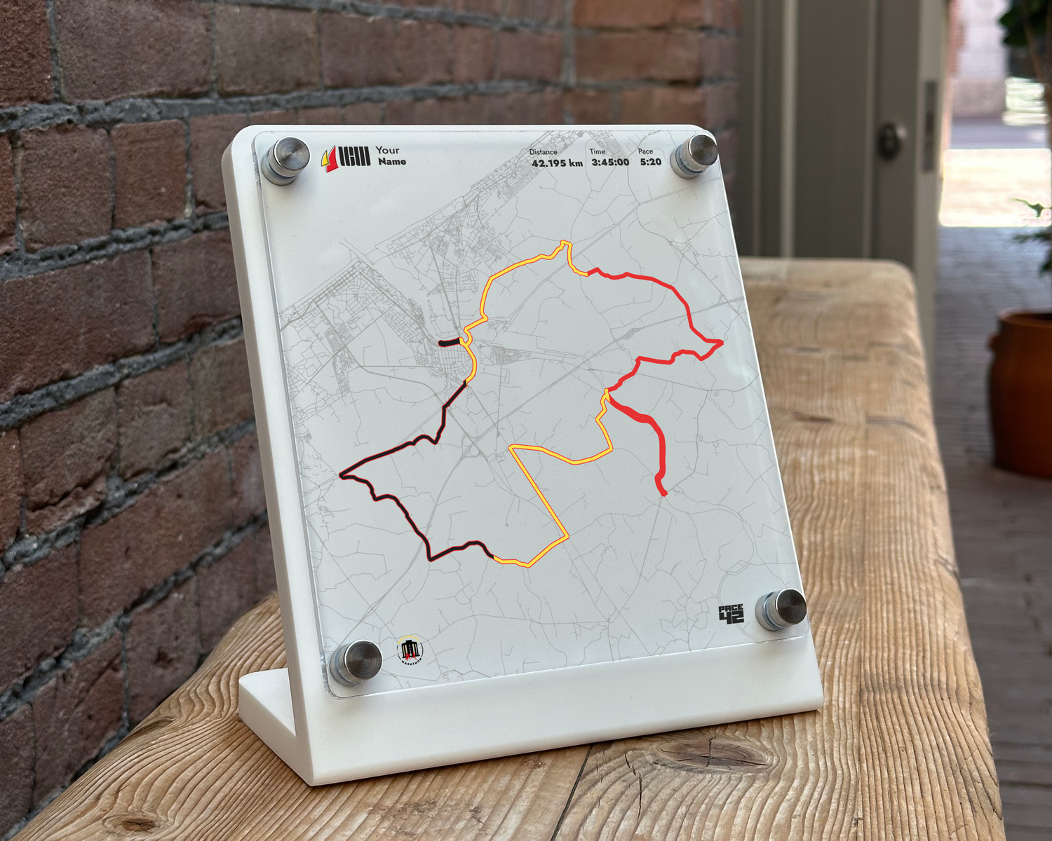 A white acrylic fitness board with a printed map and colored workout lines, displayed on a wooden table in front of a brick wall.
