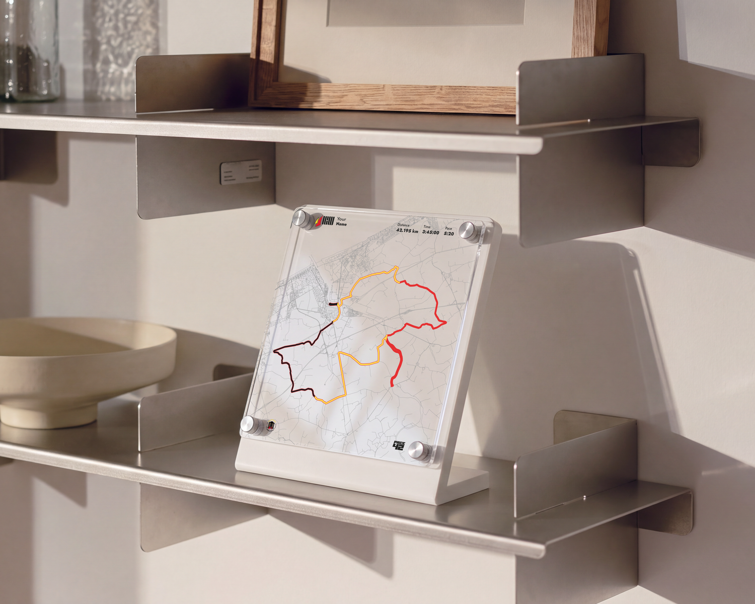 A modern wall-mounted floating shelf system in a warm beige/taupe finish, styled with a ceramic bowl, a small framed print, and a glass container against a beige wall.